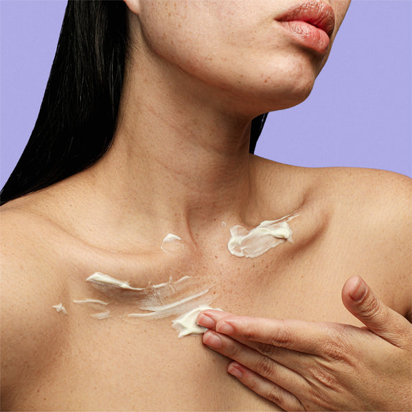 A hand smoothing cream from the frank body Always On My Niacina-mind Kit in white streaks across a bare neck and chest on a lilac background.