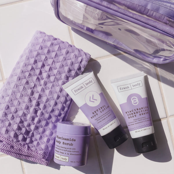 Flat lay of the frank body Always On My Niacina-mind Kit with the body scrub tub, retinol lotion, correcting serum, clear bag and lilac waffle towel on tiles.
