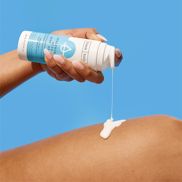 A hand tipping a bottle of frank body Hyaluronic Acid Body Serum, the cream serum dripping onto a leg below.