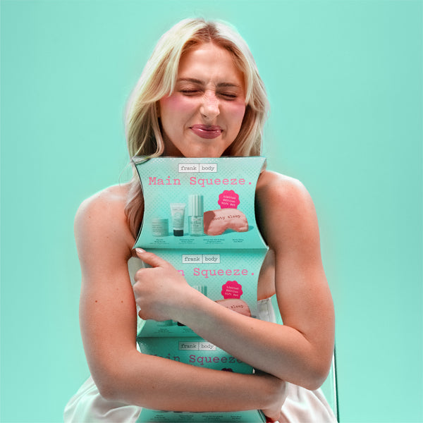A blonde woman in a white dress hugs a stack of Fresh & Body Main Squeeze hand towels against a solid mint green background.