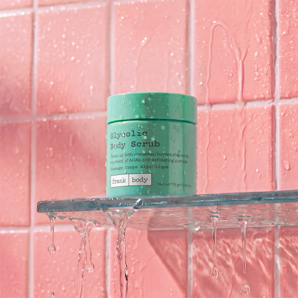A jar of Frank Body Glycolic Body Scrub in mint green, placed on a glass shelf with water dripping from it against a pink tiled background.