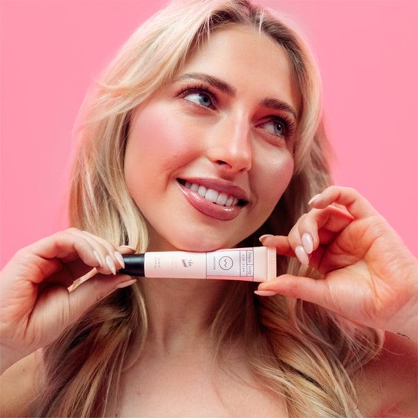A smiling blonde woman holds a small cosmetic tube against a solid pink background. The tube appears to be a liquid beauty product, shown close up in a clean, studio-style lifestyle shot.