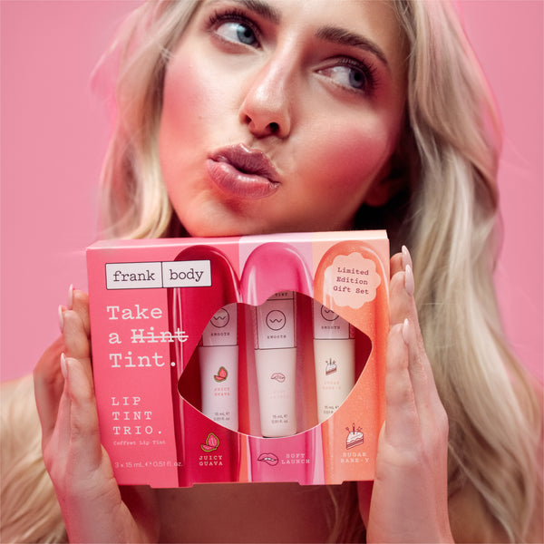 A woman with blonde hair holds a pink gift box containing three lip tint tubes. The box is branded 'frank body' and reads 'Take a Hint, Tint.