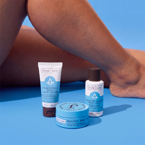 The frank body Thirst Busters Kit body serum, barrier cream and body wash set beside a model's bare legs on a blue background.