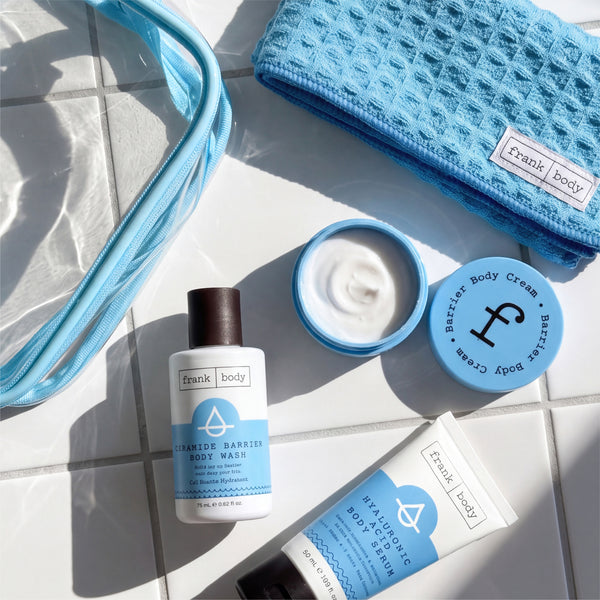 Flat lay of the frank body Thirst Busters Kit with the open barrier body cream jar, body wash, body serum, clear bag and blue waffle towel on white tiles.