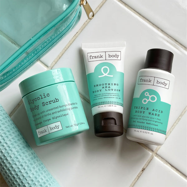Flat lay of the frank body Triple Acid Drop Kit with the scrub tub, lotion tube, body wash, clear bag and mint waffle towel on white tiles.