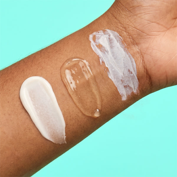 Three swatches from the frank body Triple Acid Drop Kit on a forearm: a white lotion, a clear gel and a grainy scrub, on a mint background.