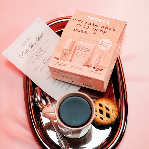 Frank body Full Body Boost kit displayed on a metallic tray with a cup of coffee, a cookie, and a printed insert card against a pink background.
