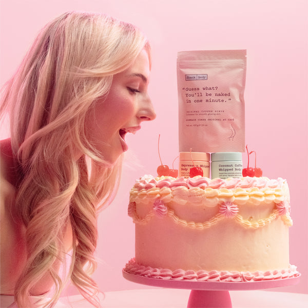 A woman with long blonde hair leans in toward a pink frosted cake on a stand, with a pink bag and two small candles placed on top of the cake against a solid pink background.