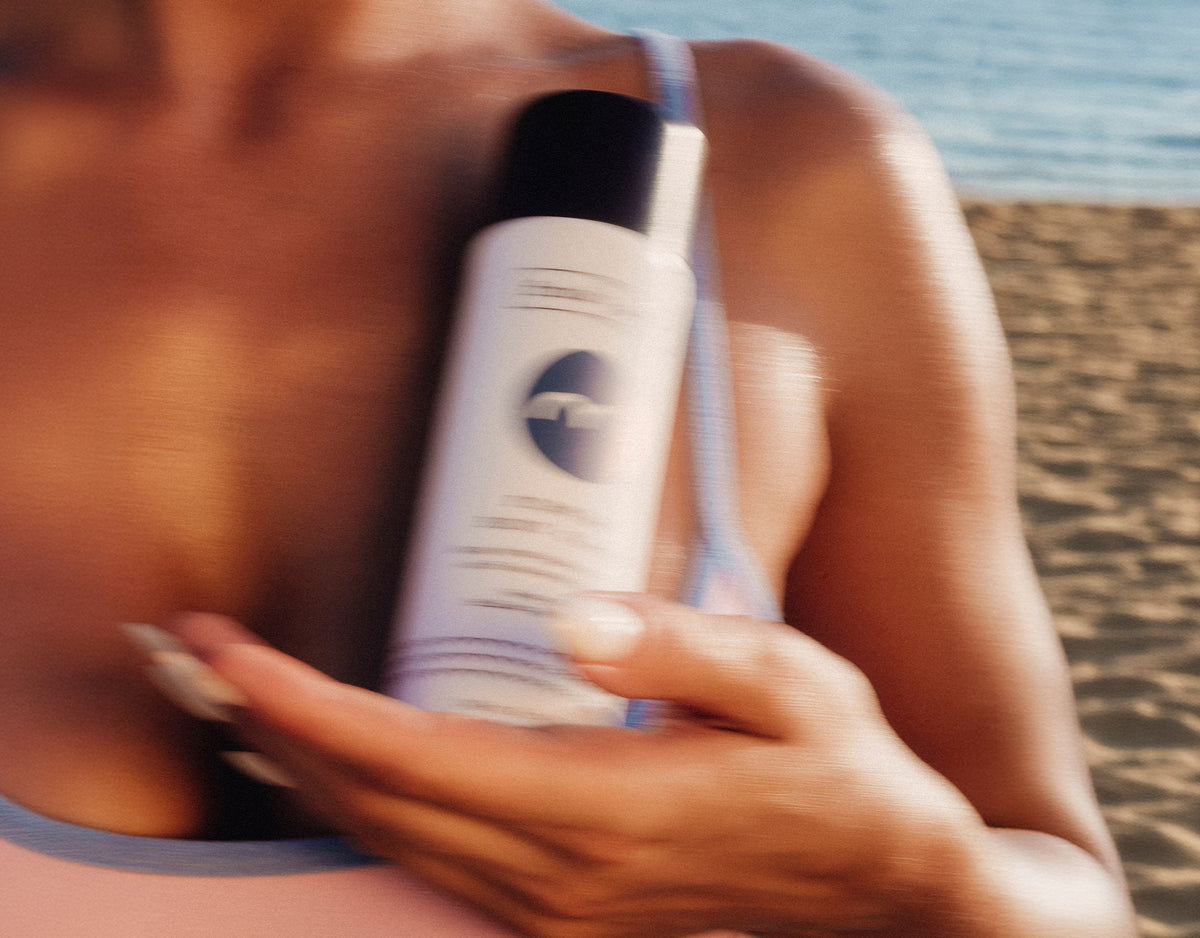 A person at the beach holding a white firming body glaze bottle with a black cap against their chest.